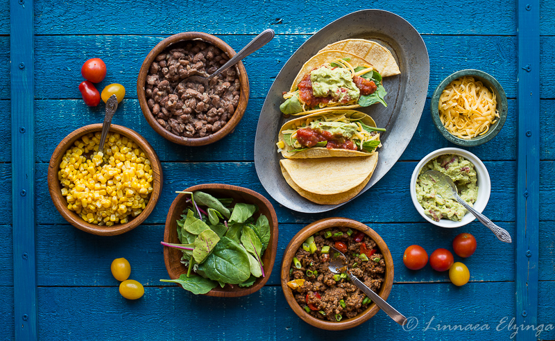 Chipotle-style buffet spread with taco meat, corn, tortillas, beans, guacamole, cheese, and greens. 